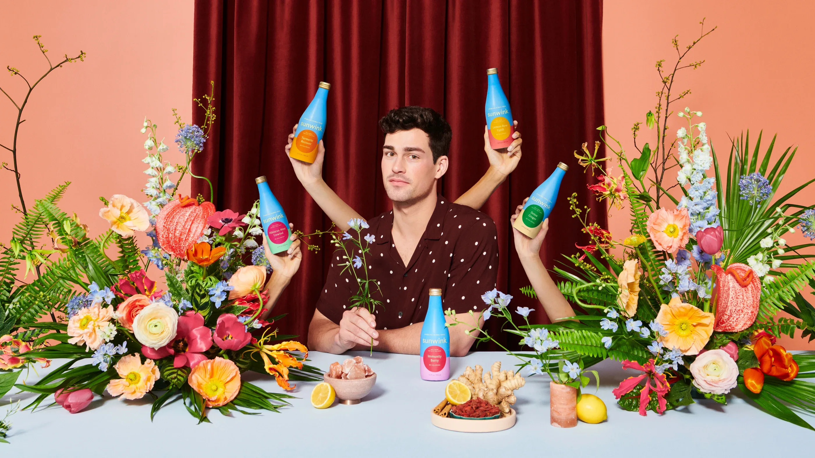 A man at a table of flowers, four arms around him raising Sunwink bottles, against a red curtain
