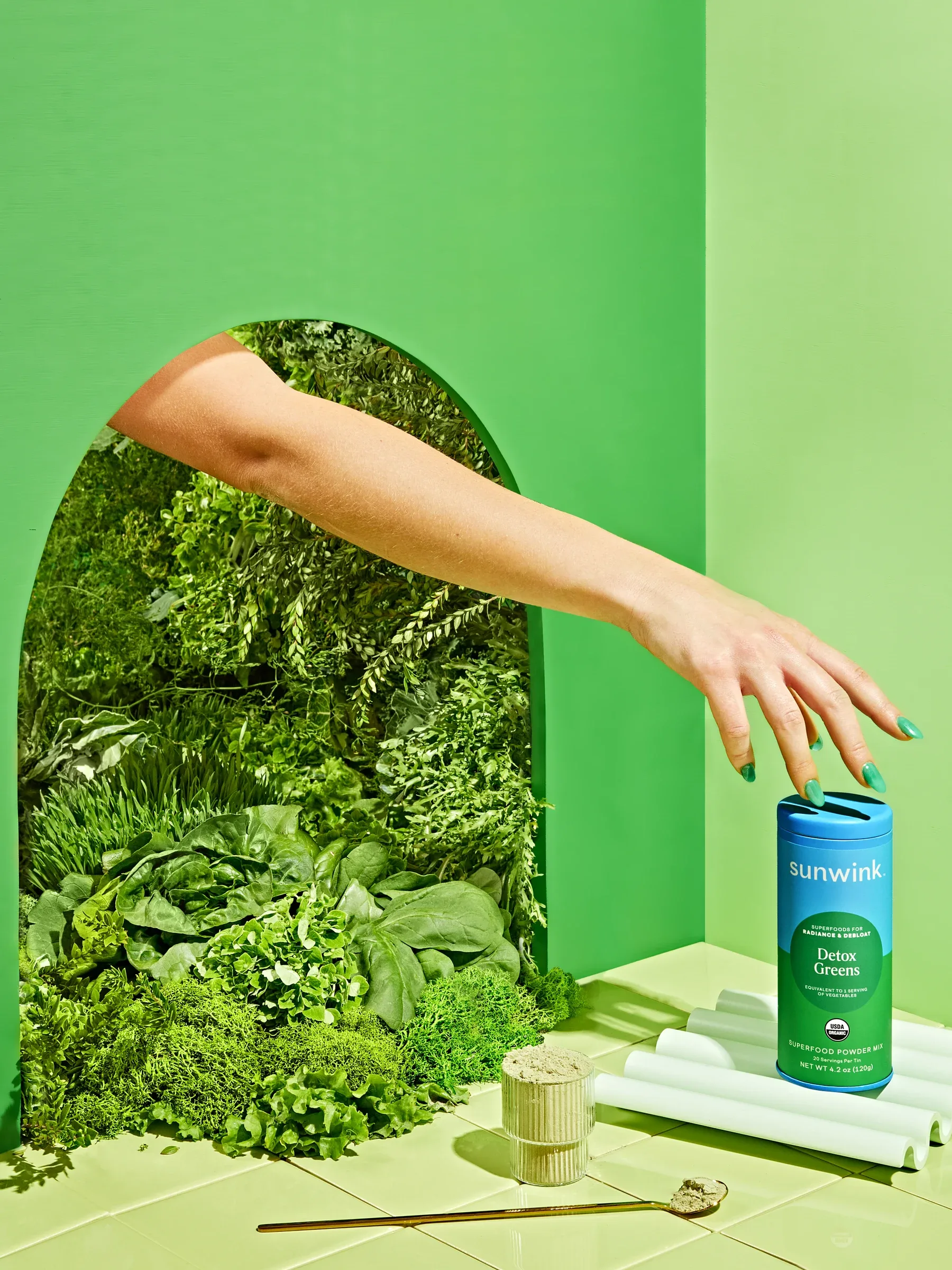 An arm reaching through an arch of leaves in a green set to place a tin of Detox Greens