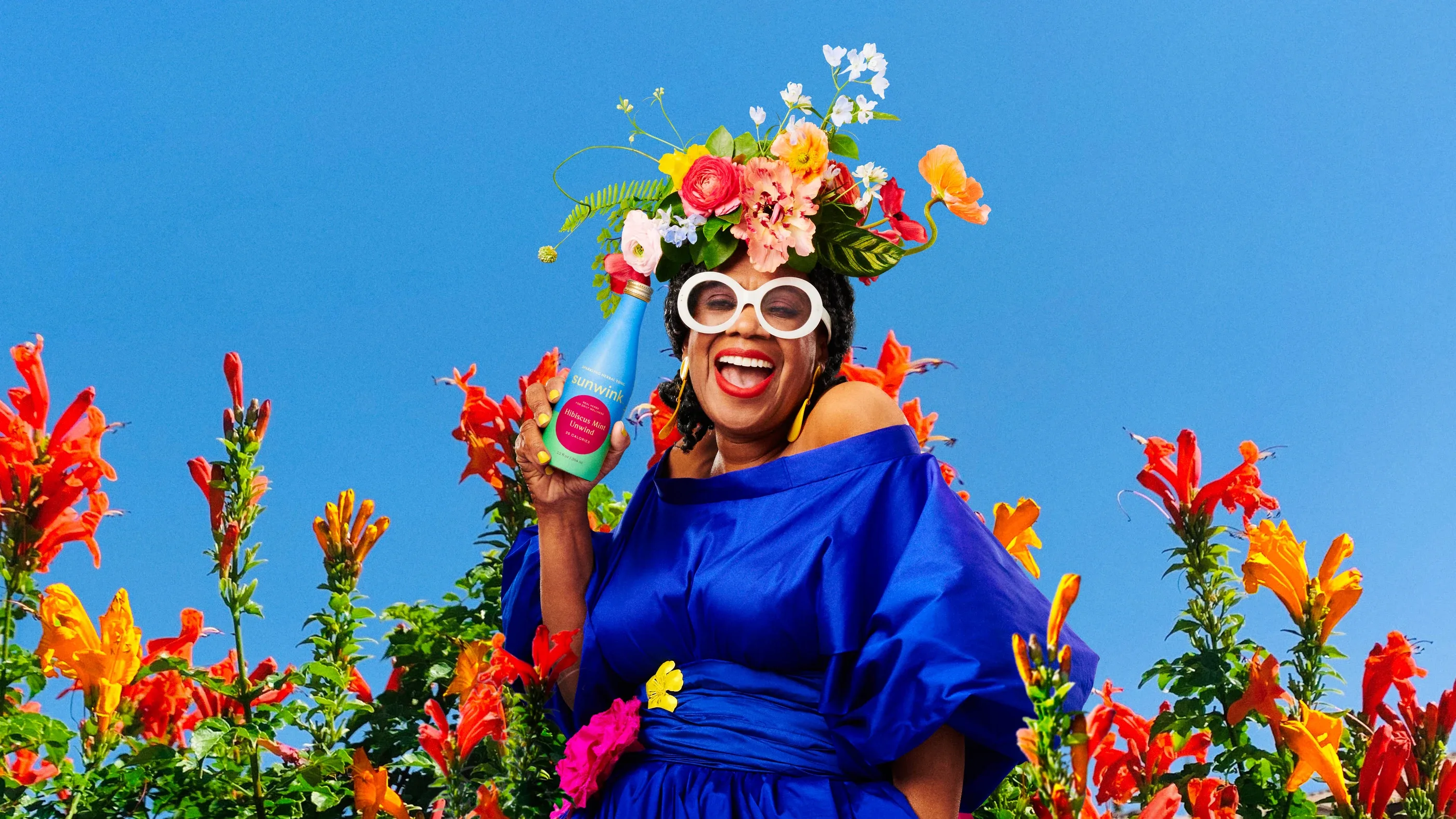 A woman in a blue dress and a crown of flowers laughing, holding a Sunwink bottle, among red flowers against a blue sky